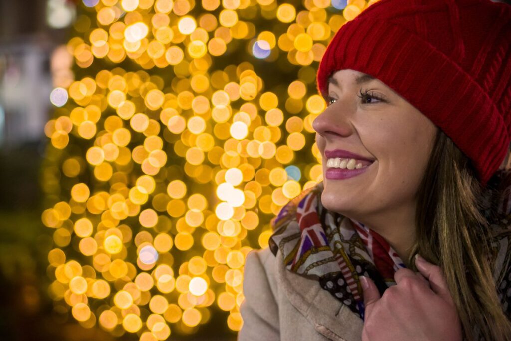 Smiling woman in red winter hat enjoys holiday lights, ready to sparkle this season.