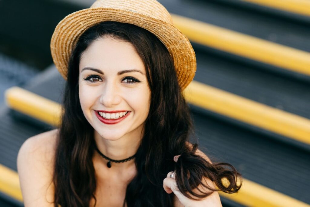 Smiling woman with healthy teeth wearing a straw hat outdoors.