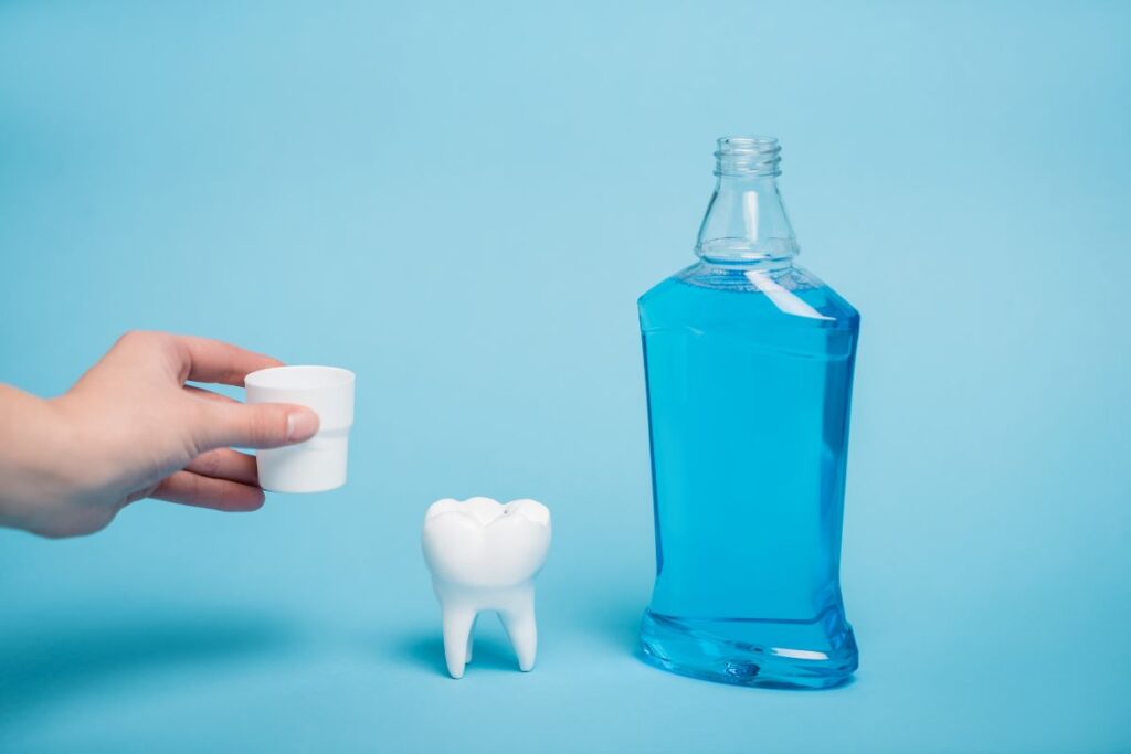 Hand holding cup next to tooth model and blue mouthwash bottle on blue background.